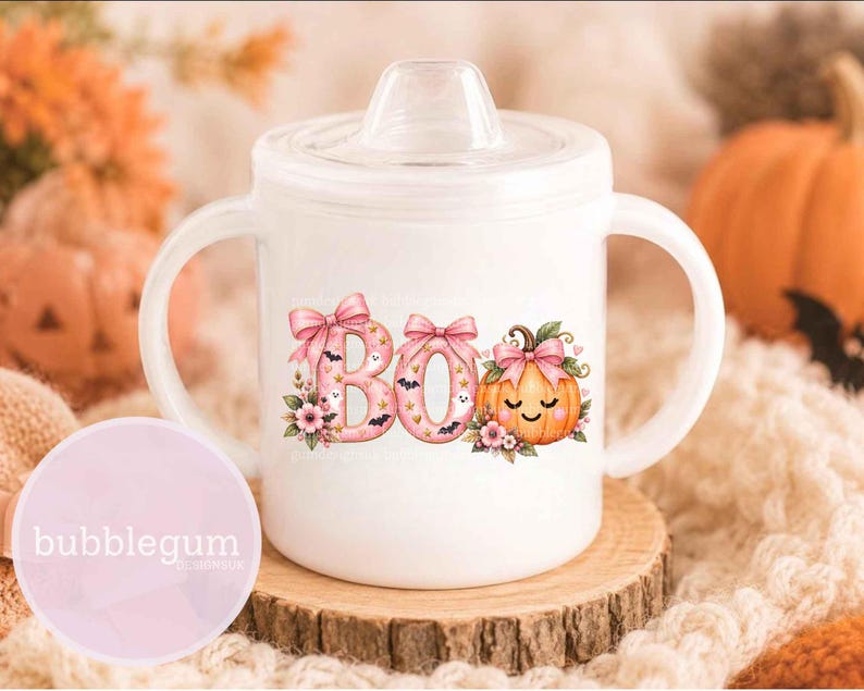 May include: A white sippy cup with two handles and a clear lid, featuring a pink graphic that spells BOO with bows, small ghosts, bats, and a smiling pumpkin with a pink bow, decorated with floral accents and gold stars on the front surface.