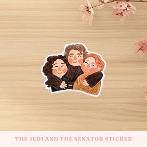 May include: A sticker depicting three cartoon characters hugging, with happy expressions and dark hair. The sticker is on a light wood surface, with the text "THE JEDI AND THE SENATOR STICKER". Pink flowers are in the background.