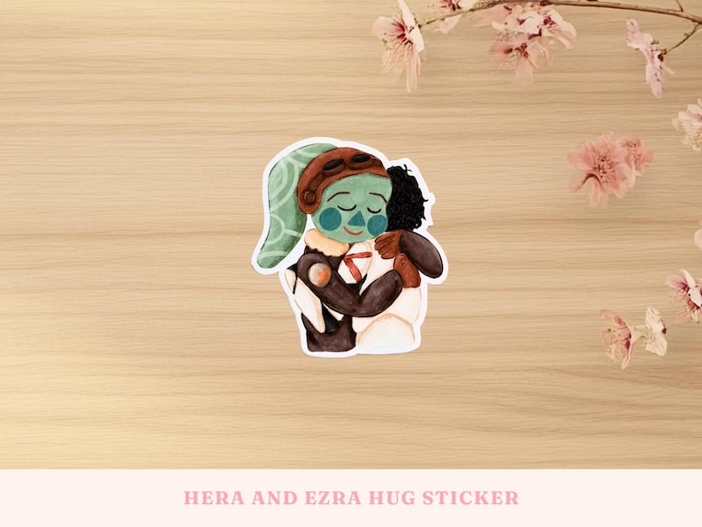 May include: A sticker featuring a watercolor illustration of two characters embracing. One character has green skin, a brown aviator cap, and a white and brown outfit. The other character has black hair and a brown outfit. The sticker's text reads "HERA AND EZRA HUG STICKER."