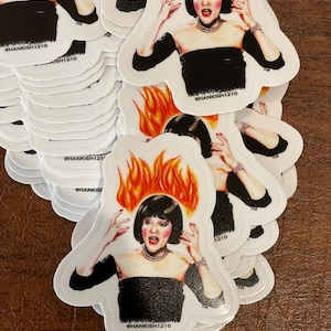 May include: A stack of white stickers featuring a woman with black hair and a black dress, with flames on her head. The woman has a surprised expression and is looking upwards. The stickers have the text "@HANKISH1210" on them.