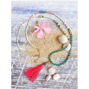 May include: A green and pink beaded necklace with a pink tassel. The necklace is on a wooden surface with white stones and a pink flower.