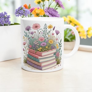 May include: White ceramic mug featuring a stack of books with colorful spines, topped with a vibrant arrangement of flowers in pink, yellow, and purple. The mug has a curved handle and is set on a light wooden surface.