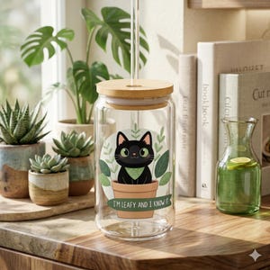 Puede incluir: Un tarro de cristal transparente con tapa de bambú y pajita de cristal. El tarro presenta un gato negro de dibujos animados en una maceta con el texto "I'M LEAFY AND I KNOW IT". El tarro está rodeado de plantas y libros.