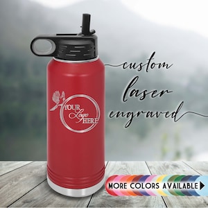 May include: Red stainless steel water bottle with a black lid and a carrying handle. The bottle has a straw lid and is engraved with the words "Your Logo Here". The text "custom laser engraved" is written in cursive above the bottle. The text "MORE COLORS AVAILABLE" is written below the bottle.
