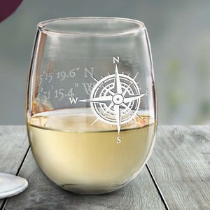 May include: A stemless wine glass filled with white wine. The glass is etched with a compass rose design and coordinates. The glass sits on a wooden surface. The wine glass is clear.