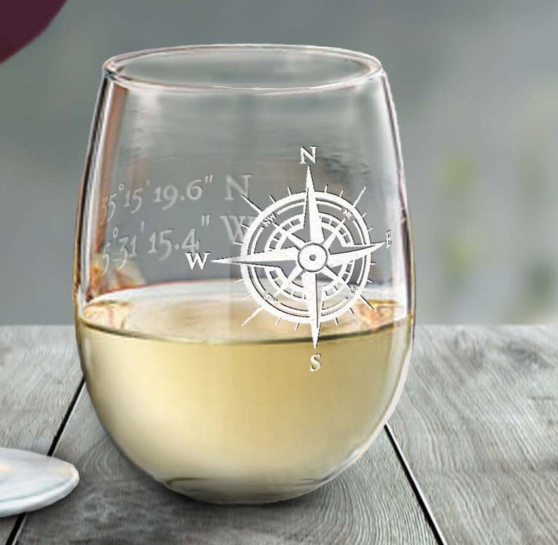 May include: A stemless wine glass filled with white wine. The glass is etched with a compass rose design and coordinates. The glass sits on a wooden surface. The wine glass is clear.