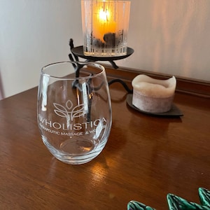 May include: A clear, stemless wine glass with the text "Wholistica Therapeutic Massage & Wellness" in white. The glass sits on a dark wooden surface, next to a lit candle in a glass holder and a smaller candle.