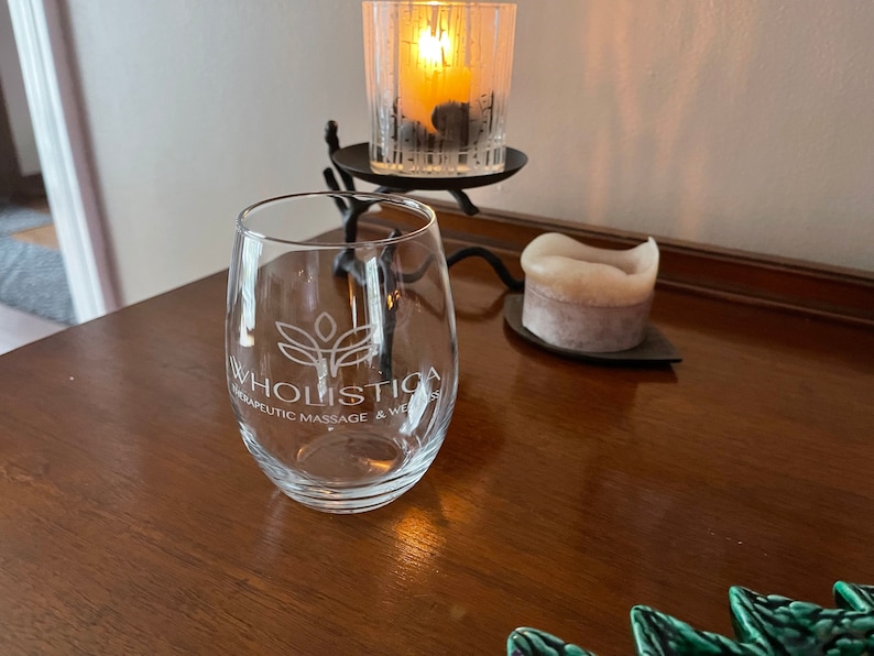 May include: A clear, stemless wine glass with the text "Wholistica Therapeutic Massage & Wellness" in white. The glass sits on a dark wooden surface, next to a lit candle in a glass holder and a smaller candle.