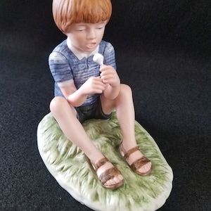 Puede incluir: Una figurita de porcelana de un niño pequeño sentado en un parche de hierba, con una camisa a rayas azul y blanca, sandalias marrones y sosteniendo una flor blanca.