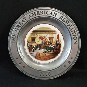 May include: A pewter plate with a white porcelain center featuring a painting of the signing of the Declaration of Independence. The plate is inscribed with "The Great American Revolution" and "1776".