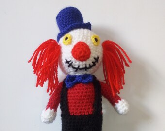May include: A crocheted clown doll with a red and white face, blue hat, red hair, and black and red clothing.