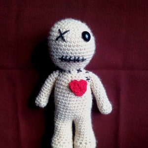 May include: A crocheted doll with a stitched-on heart, one black eye, and an X stitched on its forehead. The doll is beige and has a red heart on its chest.