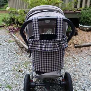 May include: A navy blue and tan plaid fabric storage bag with a ruffled top edge and two vertical straps, attached to the back of a gray stroller. The bag features a classic checkered pattern and is designed to hang securely from the stroller handle.