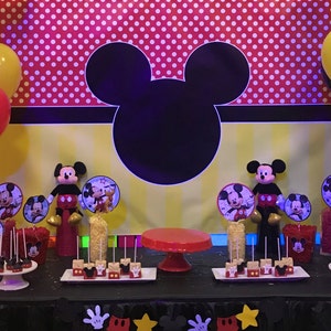May include: A Mickey Mouse themed party scene with a black Mickey silhouette, red and white polka dots, and yellow stripes as a backdrop. Red, yellow, and black balloons are scattered around. The table is decorated with treats and Mickey Mouse figurines, creating a celebratory atmosphere.