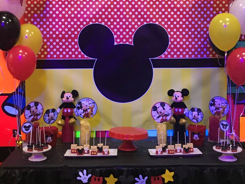 May include: A Mickey Mouse themed party scene with a black Mickey silhouette, red and white polka dots, and yellow stripes as a backdrop. Red, yellow, and black balloons are scattered around. The table is decorated with treats and Mickey Mouse figurines, creating a celebratory atmosphere.