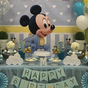 May include: A blue and yellow Mickey Mouse themed first birthday party dessert table with a blue tablecloth, white cloud-shaped cake stands, and a banner that says "Happy Birthday".