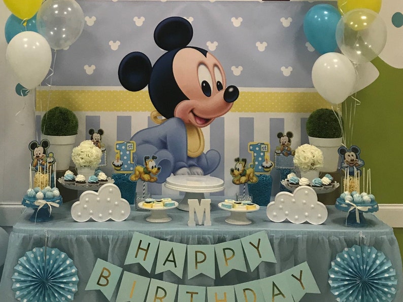 May include: A blue and yellow Mickey Mouse themed first birthday party dessert table with a blue tablecloth, white cloud-shaped cake stands, and a banner that says "Happy Birthday".