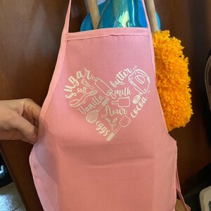以下が含まれることがあります： 白いハート型のデザインが施されたピンク色の綿のエプロン。デザインには「砂糖、塩、バニラ、バター、ミルク、小麦粉、卵、ココア」の文字と、ベーキングの材料や道具のイラストが描かれています。