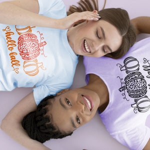 May include: Two people wearing t-shirts with a pumpkin and the text "hello fall" on them. The person on the left is wearing a light blue shirt with orange pumpkins and the person on the right is wearing a purple shirt with black pumpkins.