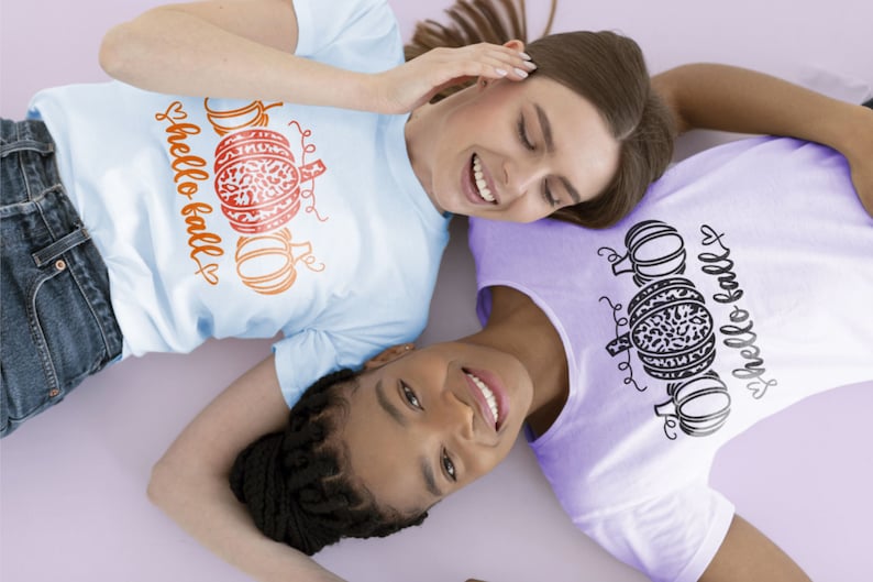 May include: Two people wearing t-shirts with a pumpkin and the text "hello fall" on them. The person on the left is wearing a light blue shirt with orange pumpkins and the person on the right is wearing a purple shirt with black pumpkins.