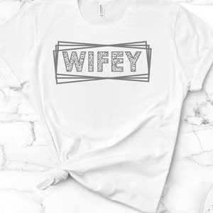 May include: White t-shirt with the word "WIFEY" printed in gray with a leopard print pattern inside the letters. The word is surrounded by a gray outlined box.