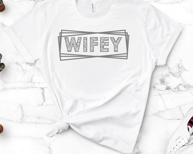 May include: White t-shirt with the word "WIFEY" printed in gray with a leopard print pattern inside the letters. The word is surrounded by a gray outlined box.