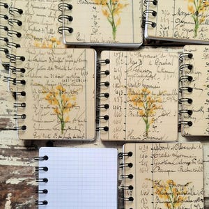 May include: Six small, spiral bound notebooks with floral designs and handwritten text. The notebooks are on a wooden surface.