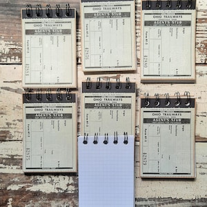 May include: Six small, vintage-style spiral notebooks with black covers and white pages. Each notebook has a printed label that reads "Ohio Trailways" and "Agent's Stub" with a form number and a number. The notebooks are arranged in two rows of three on a wooden surface.