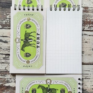 May include: Four small, white, spiral bound notebooks with green and gold labels. The labels feature a horse and rider with the text "TURF" and "Flor de Tabacos Especiales" in Spanish. The notebooks are on a wooden surface.