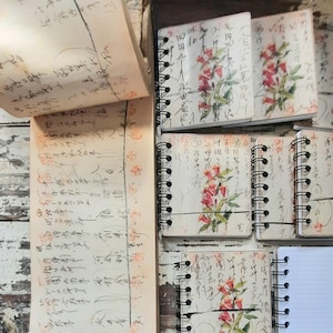 May include: A collection of spiral-bound notebooks with floral designs and handwritten script. The covers feature red and pink flowers with green leaves. The pages inside are filled with handwritten text and lines. The notebooks are arranged on a distressed wooden surface.