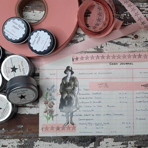 May include: A collection of pink Tele-type paper tins, rolls of pink ribbon with star patterns, and a vintage ledger. The tins have black lids and white labels. The ledger features handwritten entries and a woman illustration.