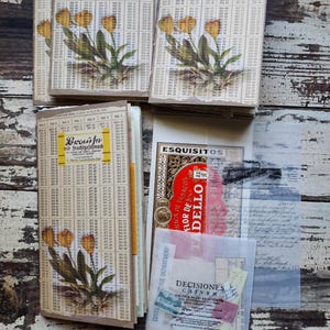 May include: A collection of vintage paper ephemera, including ledger pages, a tobacco label, and various small paper scraps. The ledger pages feature floral decals of yellow tulips. The items are arranged on a distressed white wooden surface.