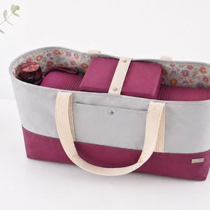 May include: A grey and burgundy tote bag with cream handles, containing yoga mats and blocks. The interior has a floral pattern. The bag is designed for carrying yoga equipment and accessories.