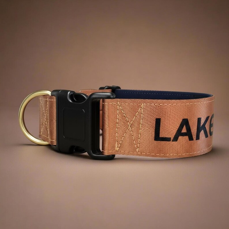 May include: A tan nylon dog collar featuring a black plastic side-release buckle, a gold-toned metal D-ring, and the text LAK printed in black on the outer strap. The interior of the collar is lined with a dark navy blue fabric material.