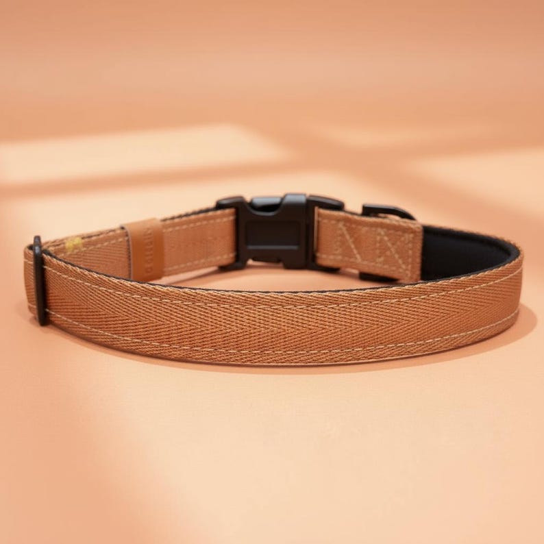 May include: A tan-colored pet collar featuring a durable herringbone-patterned fabric strap, a black plastic side-release buckle, a matching black metal D-ring, and a small tan leather-like accent tab stitched onto the side of the webbing.