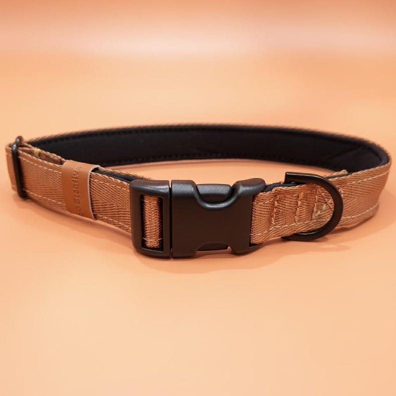May include: A tan-colored pet collar featuring a black plastic side-release buckle, a black metal D-ring, and a black interior lining. The exterior fabric has a visible diagonal weave pattern and a small tan tag with text attached to the side.