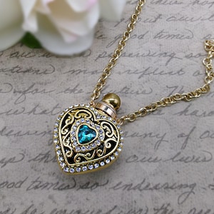 May include: A gold-colored heart-shaped pendant necklace with a teal-colored gemstone at the center. The pendant is adorned with clear crystals and intricate filigree detailing. The necklace has a delicate chain.