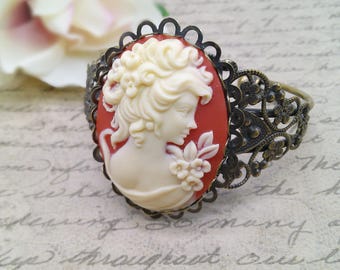May include: A vintage-style bracelet with a cameo featuring a woman's profile. The cameo is set in a red resin and surrounded by an ornate bronze filigree design.