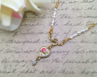 May include: A delicate gold-toned necklace featuring a heart-shaped pendant with a pink rose design. The necklace includes a small, sparkling crystal and is accented with white pearls. The background is a vintage-style paper with handwritten text.