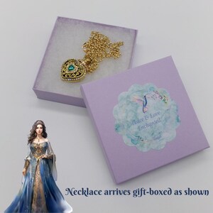 May include: A gold-colored heart-shaped pendant necklace with a teal-colored gem and intricate detailing, presented in a lavender gift box. The necklace is on a gold chain. The box has a sticker that says "Peace & Love Enchanted".