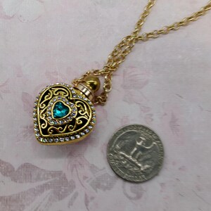 May include: A gold-colored heart-shaped pendant necklace with a teal-colored gemstone in the center. The pendant is adorned with clear crystals and intricate filigree detailing. The necklace is displayed on a pink floral background.