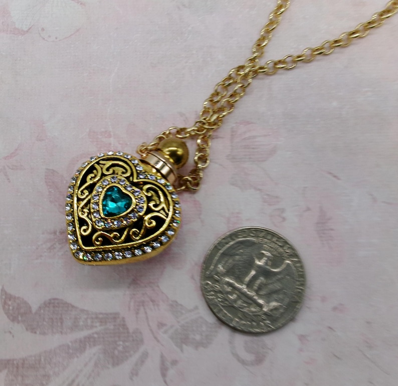 May include: A gold-colored heart-shaped pendant necklace with a teal-colored gemstone in the center. The pendant is adorned with clear crystals and intricate filigree detailing. The necklace is displayed on a pink floral background.