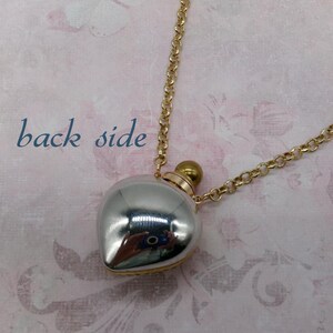 May include: A silver-colored, heart-shaped pendant necklace with a gold-colored chain. The pendant has a reflective surface and a small gold-colored cap. The words "back side" are printed in blue on the background.