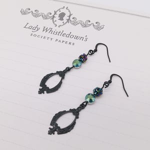 May include: Black drop earrings with an ornate, oval-shaped design. Each earring features iridescent beads and a small cluster of dark, sparkling beads. The earrings are displayed on a paper with the text "Lady Whistledown's Society Papers."