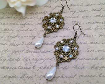 May include: A pair of dangle earrings featuring an antique bronze-toned filigree circular frame set with a central clear rhinestone and smaller surrounding crystals, finished with a single white teardrop-shaped faux pearl bead hanging below.