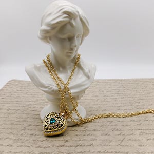 May include: A gold-colored heart-shaped pendant necklace with a teal-colored gemstone center and clear crystal accents. The pendant hangs from a long, delicate gold chain. The necklace is displayed on a white bust.