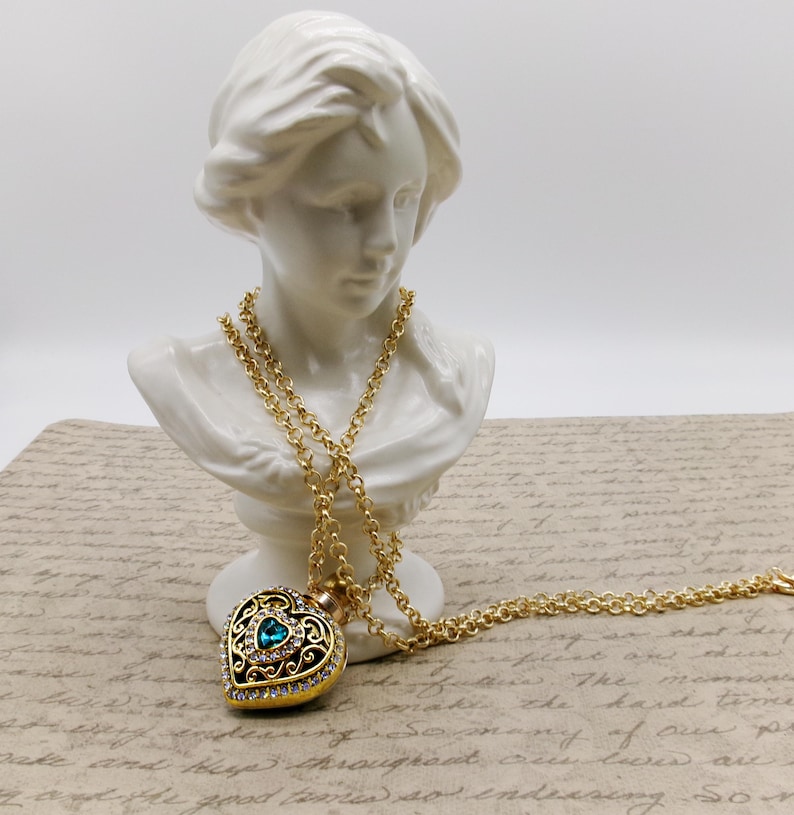 May include: A gold-colored heart-shaped pendant necklace with a teal-colored gemstone center and clear crystal accents. The pendant hangs from a long, delicate gold chain. The necklace is displayed on a white bust.