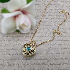 May include: Gold-toned heart-shaped pendant necklace with a turquoise center stone and clear crystal accents. The pendant has an ornate filigree design and a small bottle top. A white flower is in the background.