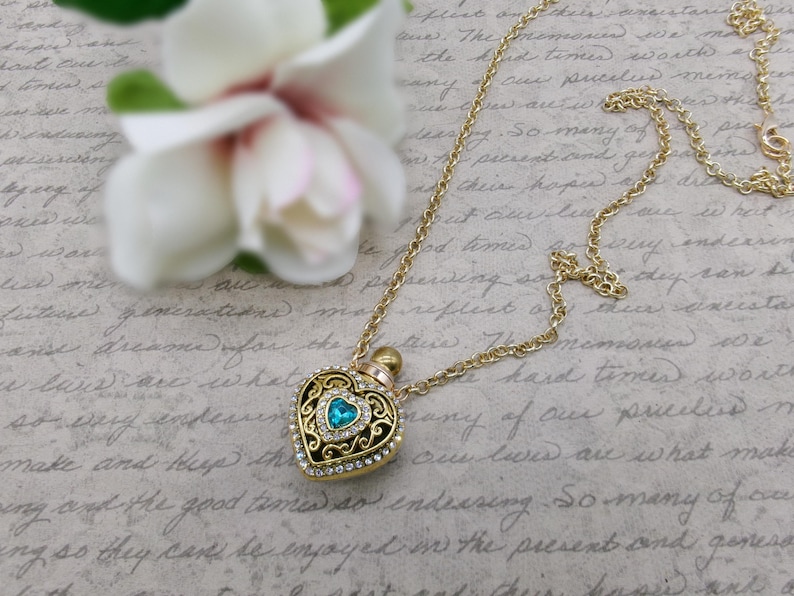 May include: Gold-toned heart-shaped pendant necklace with a turquoise center stone and clear crystal accents. The pendant has an ornate filigree design and a small bottle top. A white flower is in the background.