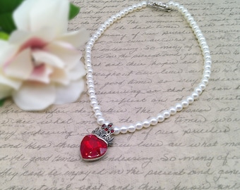 May include: A pearl necklace with a red heart pendant. The heart is adorned with a silver crown and red jewels. The necklace is on a white background with handwritten text.